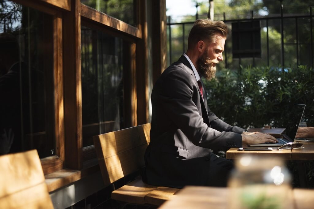 A bearded man in a suit works on a laptop at an outdoor cafe table