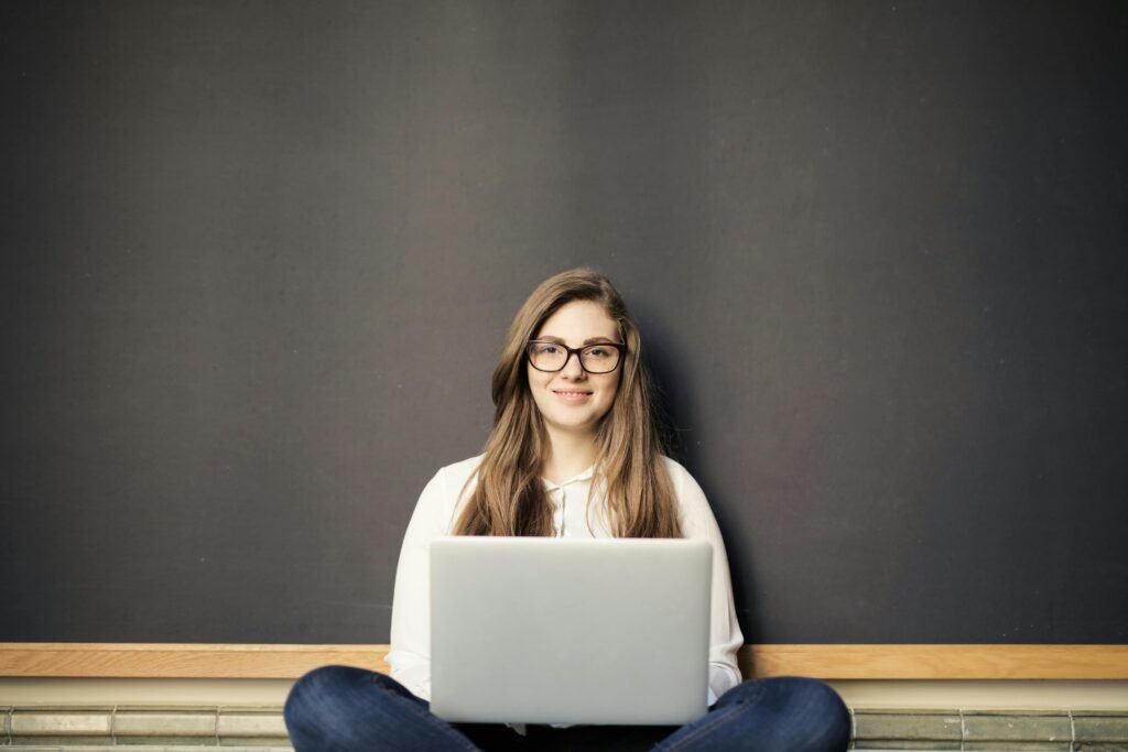 A young woman with glasses smiles while working on a laptop in front of a blackboard, demonstrating the use of ai for images