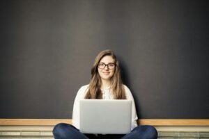 A young woman with glasses smiles while working on a laptop in front of a blackboard, demonstrating the use of ai for images