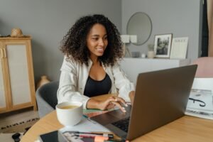 A Black woman with curly hair smiles while working on a laptop at a round wooden table, with a cup of coffee and papers nearby