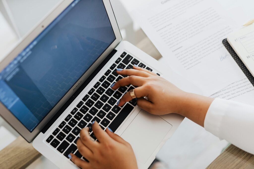 Hands with blue nail polish type on a MacBook Air next to documents and a planner, useful for alt text generation