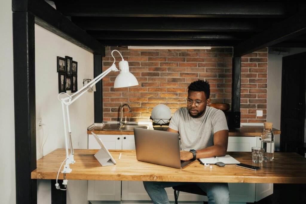 A Black man wearing glasses works on a laptop at a wooden desk, with a tablet and a glass of water nearby