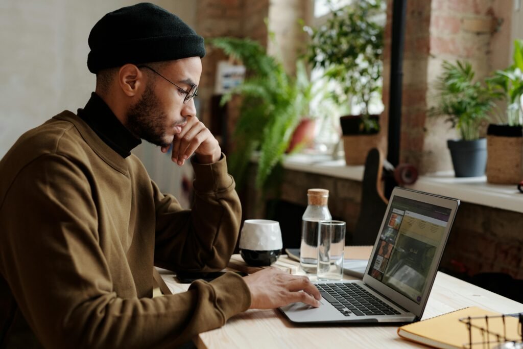 A man wearing a black beanie and glasses works on his laptop, with plants in the background