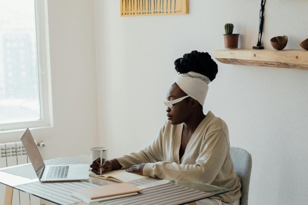 A Black woman wearing glasses and a headwrap works on her laptop and notebook at a glass table