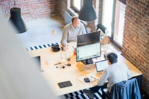 Two men work on laptops at a wooden desk in a loft-style office with exposed brick walls