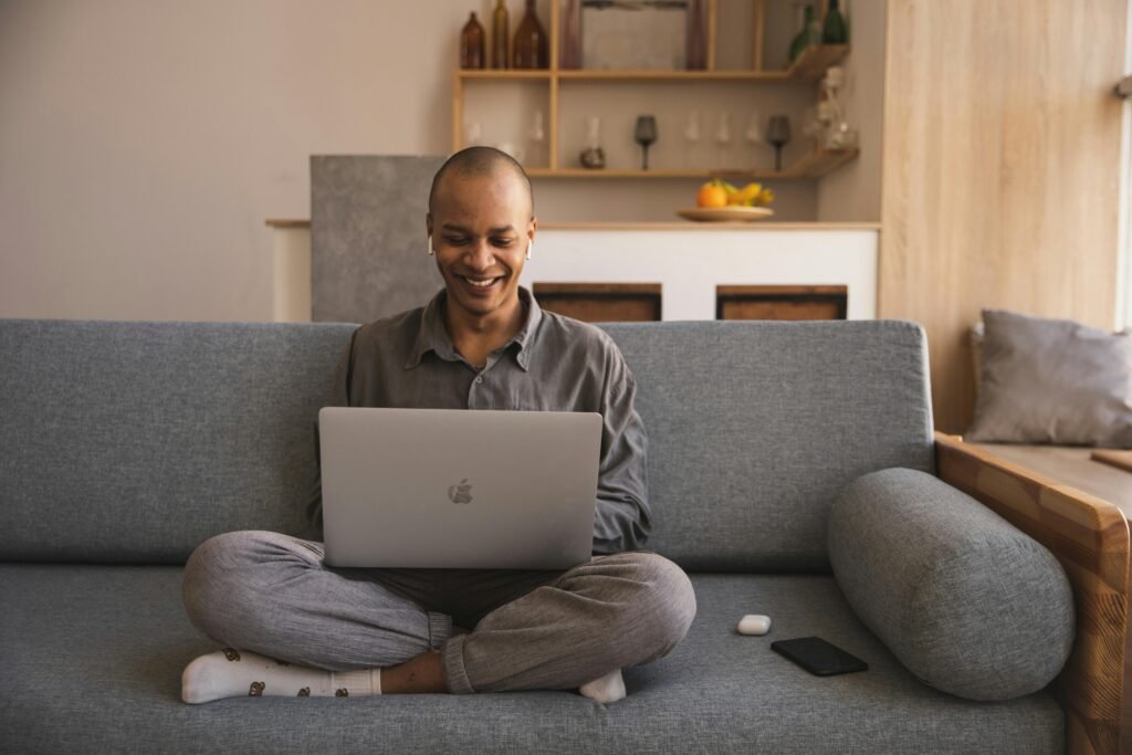 A smiling person sits cross-legged on a couch, working on a laptop, with a phone and earbuds nearby