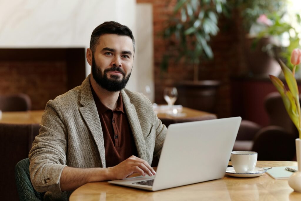 A man with a beard works on a laptop at a table with a coffee cup, near a vase with a tulip